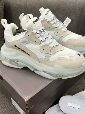 Men's luxury white Balenciaga Triple S sneakers with beige accents and distinctive clear transparent soles resting on a grey sofa.