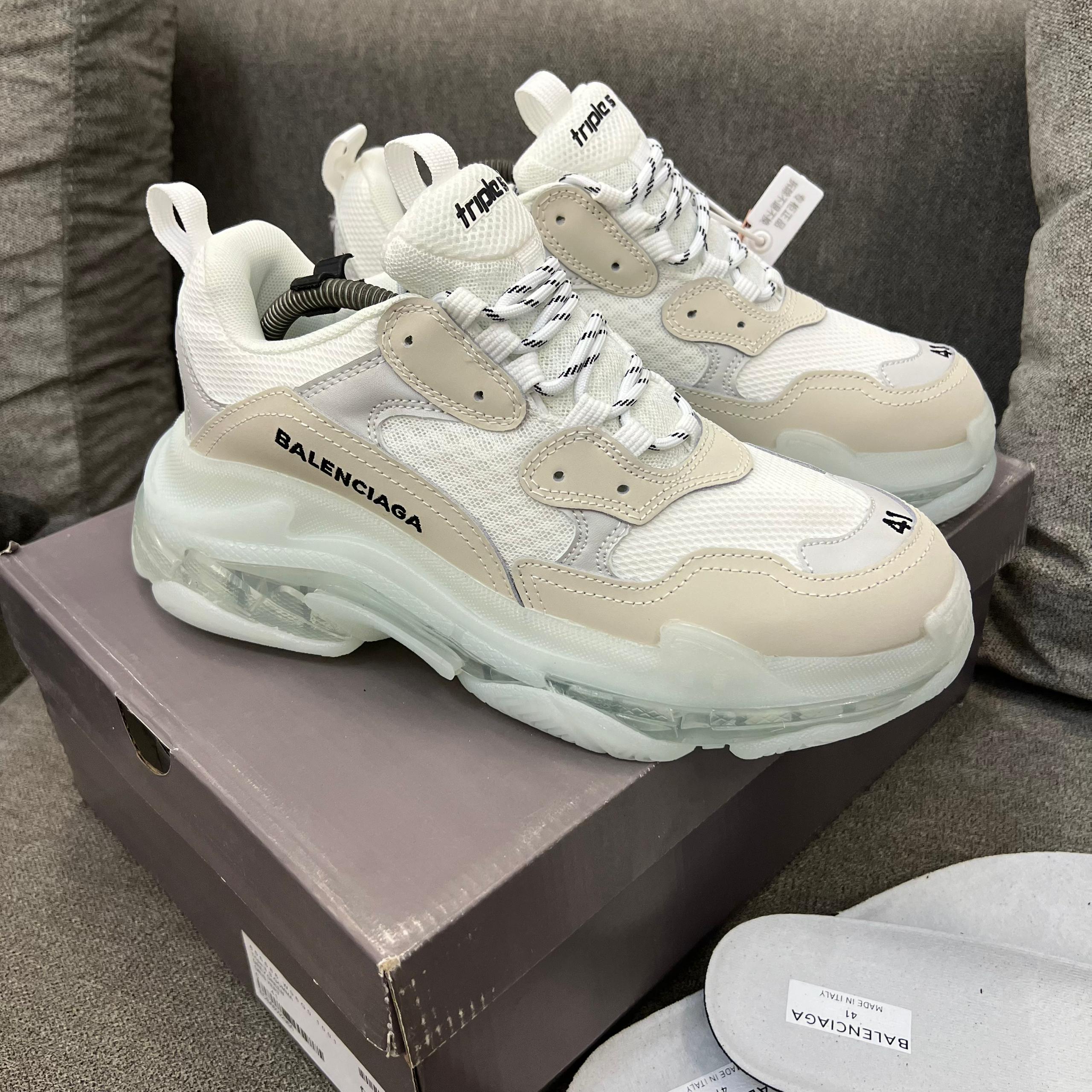 Iconic Balenciaga Triple S sneakers Men's luxury white Balenciaga Triple S sneakers with beige accents and distinctive clear transparent soles resting on a grey sofa.