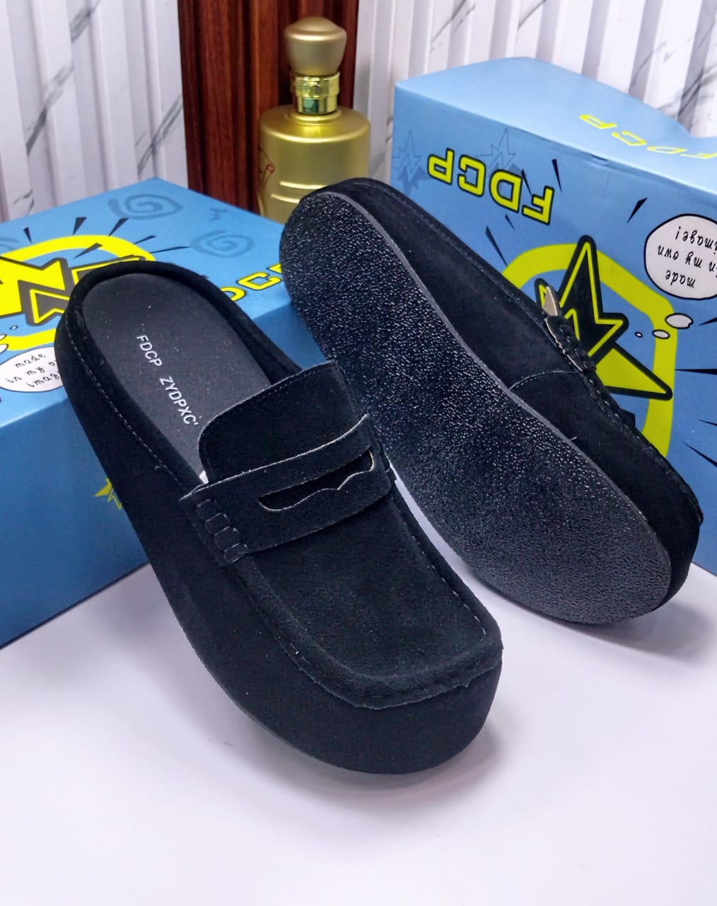FDCP taupe suede penny loafer mules FDCP men's taupe suede penny loafer mules with braided strap detail and thick rubber sole, displayed on metallic surface against stone wall background.