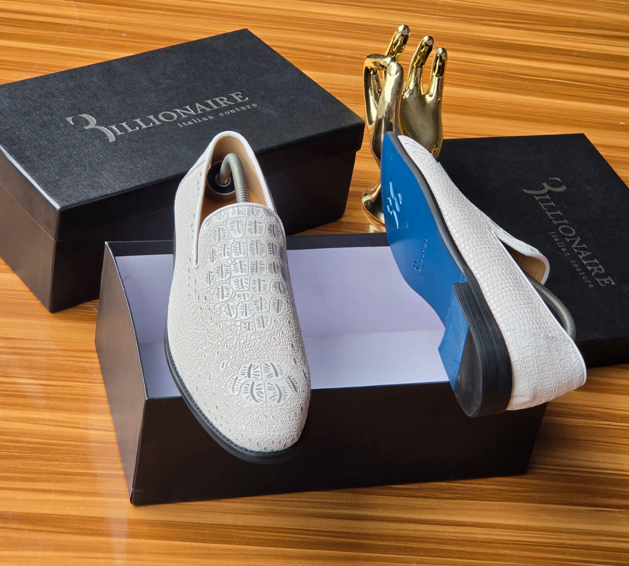 Pristine white Billionaire Italian Couture loafers Men's luxury white Billionaire Italian Couture loafers with embossed medallion pattern and blue soles resting on black branded shoeboxes.
