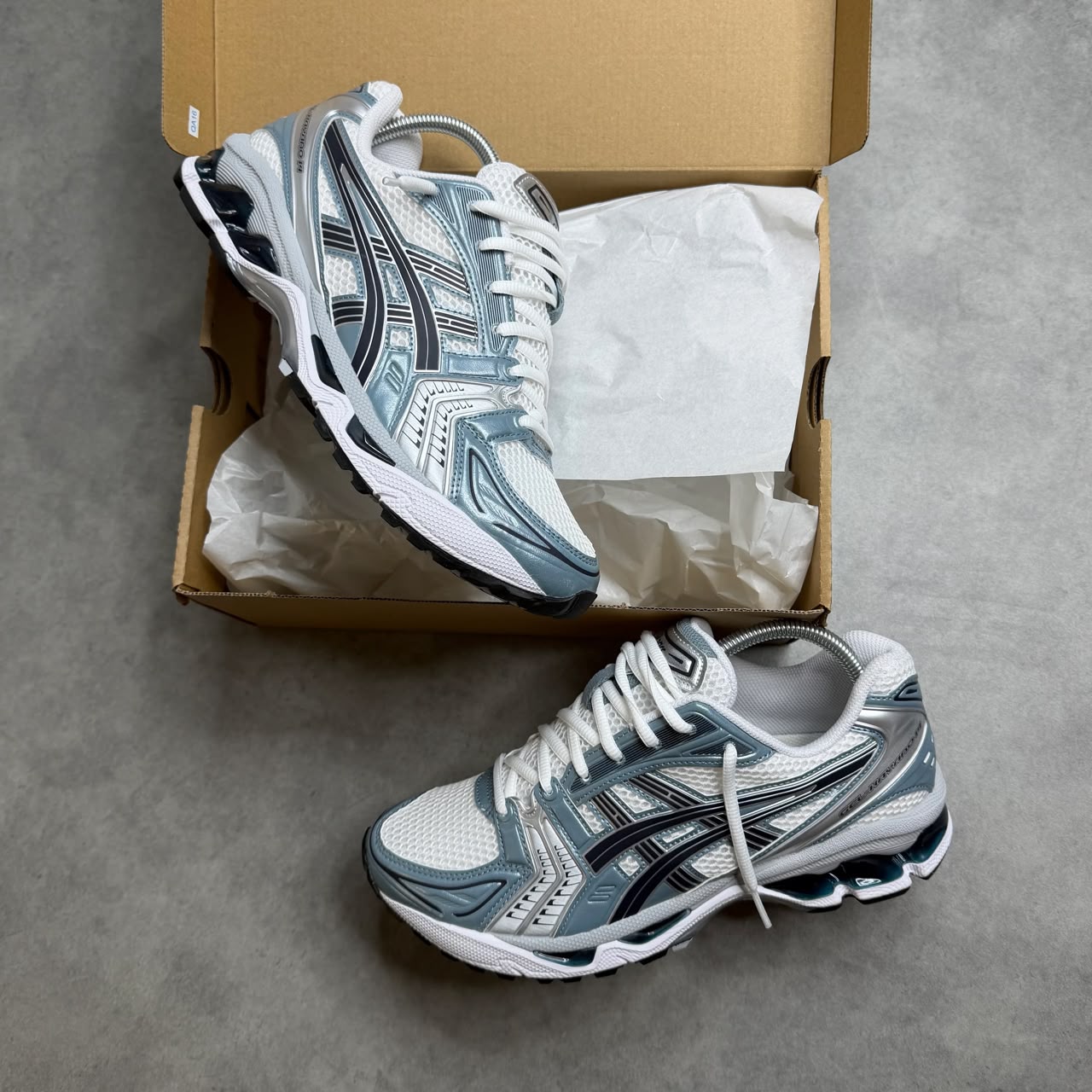 ASICS GEL-Kayano 14 sneakers in Silver/Blue ASICS GEL-Kayano 14 men's sneakers in silver, white, and blue with visible GEL cushioning units, displayed on grey concrete surface with open cardboard shoebox.