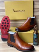 3illionaire men's cognac leather Chelsea boots with elastic side panels and red rubber sole with black studs, displayed with branded gold shoebox and decorative perfume bottle on wooden surface.
