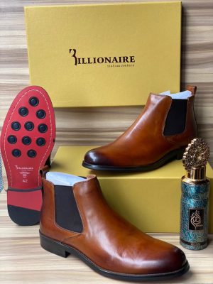 3illionaire men's cognac leather Chelsea boots with elastic side panels and red rubber sole with black studs, displayed with branded gold shoebox and decorative perfume bottle on wooden surface.