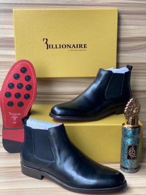3illionaire men's cognac leather Chelsea boots with elastic side panels and red rubber sole with black studs, displayed with branded gold shoebox and decorative perfume bottle on wooden surface.