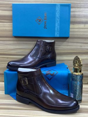 Steve Riots men's dark brown leather monk strap Chelsea boots with elastic side panels and rubber sole, displayed with branded blue shoebox and decorative perfume bottle on wooden surface.
