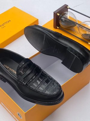 Men's chocolate brown Louis Vuitton crocodile-embossed penny loafers with metal LV hardware and rubber-injected heel, displayed with branded orange shoebox and hourglass on white surface.