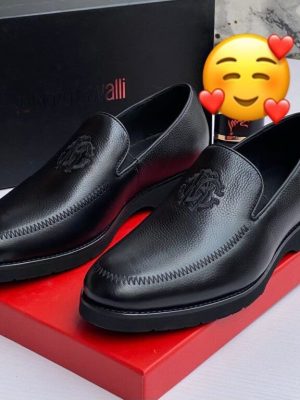 Roberto Cavalli men’s brown textured leather loafers with embossed RC monogram and rugged lug sole, displayed beside branded black box, poodle figurine, and stacked books on light wood surface.
