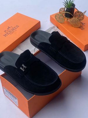 Hermès men's chocolate brown suede mule slides with silver H-hardware and black rubber sole, displayed with orange Hermès box on white surface.