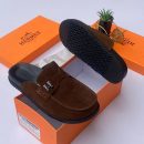 Hermès men's chocolate brown suede mule slides with silver H-hardware and black rubber sole, displayed with orange Hermès box on white surface.