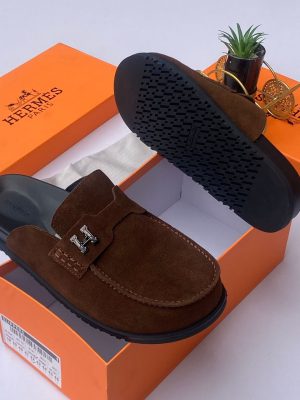 Hermès men's chocolate brown suede mule slides with silver H-hardware and black rubber sole, displayed with orange Hermès box on white surface.