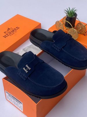 Hermès men's chocolate brown suede mule slides with silver H-hardware and black rubber sole, displayed with orange Hermès box on white surface.