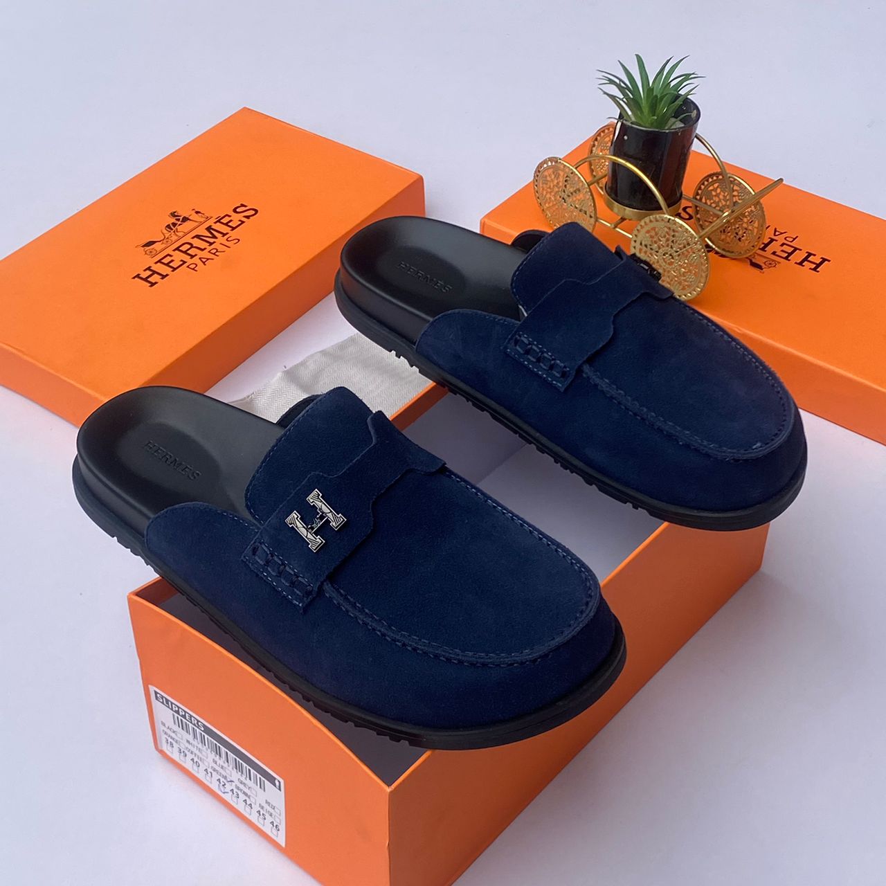 Hermès suede mule slides Hermès men's chocolate brown suede mule slides with silver H-hardware and black rubber sole, displayed with orange Hermès box on white surface.