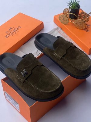 Hermès men's chocolate brown suede mule slides with silver H-hardware and black rubber sole, displayed with orange Hermès box on white surface.