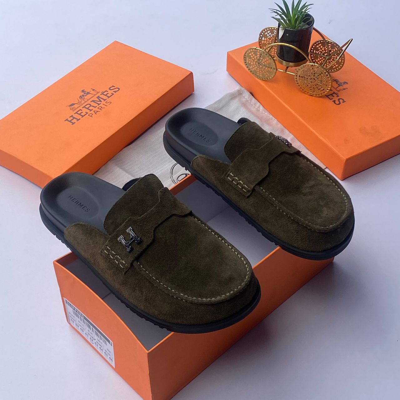 Hermès suede mule slides Hermès men's chocolate brown suede mule slides with silver H-hardware and black rubber sole, displayed with orange Hermès box on white surface.