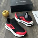 Emporio Armani men's black and red slip-on sneakers with metallic eagle logo and thick white rubber sole, displayed on a grey wood floor with a black branded shoebox and gold apple decor.