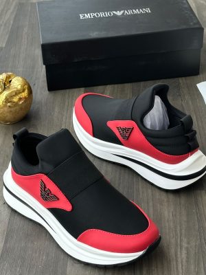 Emporio Armani men's black and red slip-on sneakers with metallic eagle logo and thick white rubber sole, displayed on a grey wood floor with a black branded shoebox and gold apple decor.