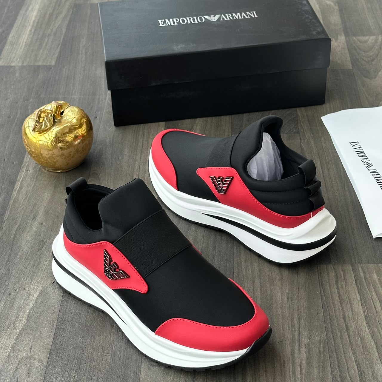 Emporio Armani black and red slip-on sneakers Emporio Armani men's black and red slip-on sneakers with metallic eagle logo and thick white rubber sole, displayed on a grey wood floor with a black branded shoebox and gold apple decor.