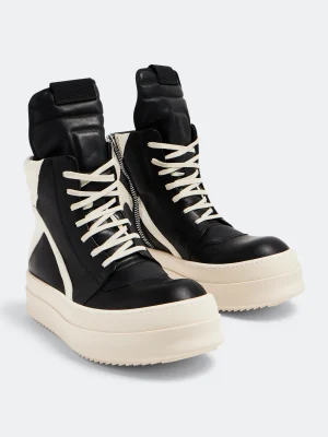 Men's black and cream high-top platform sneakers with thick white sole and side zipper, displayed on blue shoebox with fashion books and KAWS figurine in background.