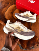 New Balance FuelCell Rebel v4 sneakers in cream and taupe mesh with gold N logo and thick white midsole, held in hand against wooden background with red NB box.