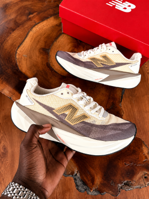 New Balance FuelCell Rebel v4 sneakers in cream and taupe mesh with gold N logo and thick white midsole, held in hand against wooden background with red NB box.