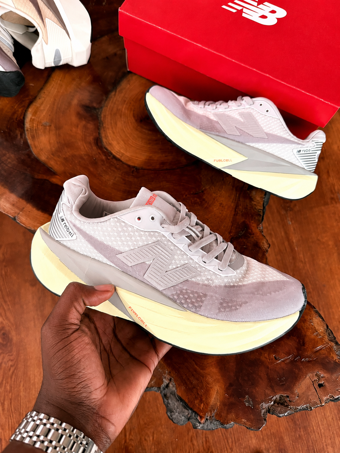 New Balance FuelCell Rebel v4 New Balance FuelCell Rebel v4 sneakers in cream and taupe mesh with gold N logo and thick white midsole, held in hand against wooden background with red NB box.