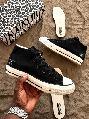 Converse x Undefeated Chuck 70 high-top sneakers in black canvas with off-white rubber sole and heel branding, held in hand with patterned shoebox and insoles visible on textured background.