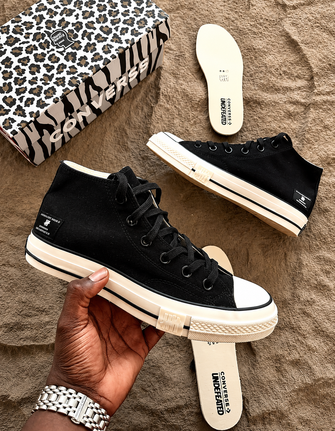 Converse x Undefeated Chuck 70 High Tops canvas Converse x Undefeated Chuck 70 high-top sneakers in black canvas with off-white rubber sole and heel branding, held in hand with patterned shoebox and insoles visible on textured background.