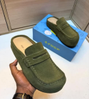 FDCP men's taupe suede penny loafer mules with braided strap detail and thick rubber sole, displayed on metallic surface against stone wall background.