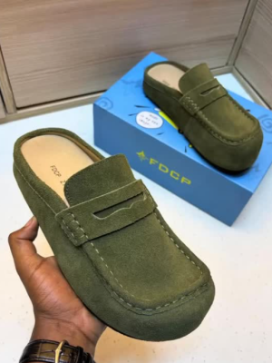 FDCP men's taupe suede penny loafer mules with braided strap detail and thick rubber sole, displayed on metallic surface against stone wall background.