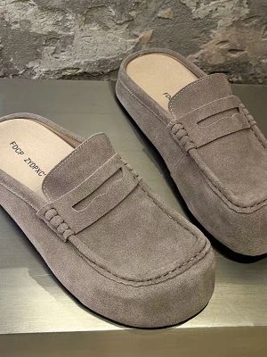 FDCP men's taupe suede penny loafer mules with braided strap detail and thick rubber sole, displayed on metallic surface against stone wall background.