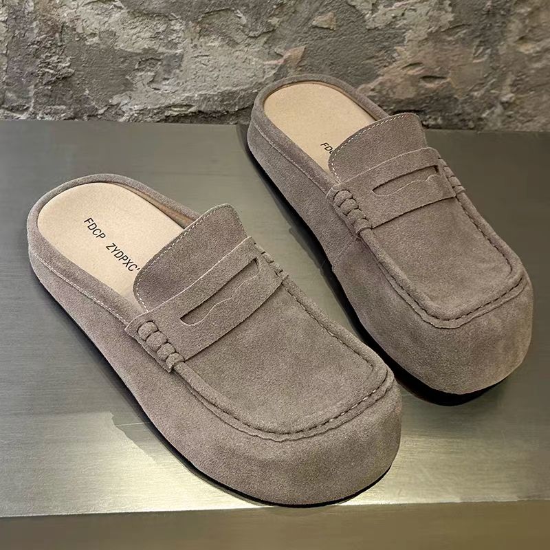 FDCP taupe suede penny loafer mules FDCP men's taupe suede penny loafer mules with braided strap detail and thick rubber sole, displayed on metallic surface against stone wall background.