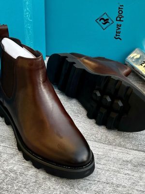 Steve Riots men’s brown burnished leather Chelsea boots with elastic sides, rugged lug sole, and gradient finish, displayed beside turquoise branded box and metallic VIP tag on textured grey surface.