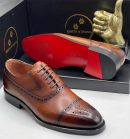 Earth Strong men’s brown leather cap-toe brogues with hand-burnished finish, perforated detailing, and signature red sole, displayed beside branded black box and golden thumbs-up sculpture on white background.