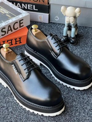 Prada men’s black leather chunky derby shoes with white lug sole and star tread pattern, displayed beside branded boxes and KAWS figurine on grey carpet background.