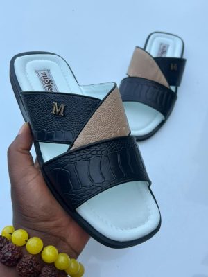 Maje men’s black and beige croc-embossed slide sandals with gold “M” hardware and white cushioned sole, held by hand beside yellow and red beaded bracelet on light blue background.