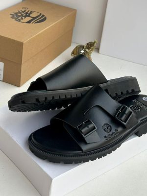 Timberland men’s black leather double-buckle slides with rugged lug sole and embossed tree logo, displayed beside branded cardboard box on white surface.