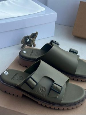 Timberland men’s black leather double-buckle slides with rugged lug sole and embossed tree logo, displayed beside branded cardboard box on white surface.