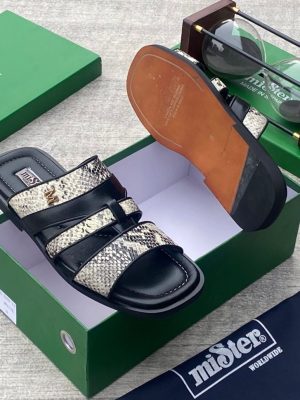 miSter men’s black and snakeskin triple-strap slides with gold “M” hardware and tan-accented sole, displayed beside green branded box and hourglass on textured surface.