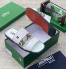 miSter men’s white and snakeskin triple-strap slides with gold “M” hardware and red-accented sole, displayed beside green branded box and hourglass on textured surface.