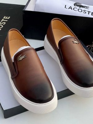 Lacoste Gazon men's slip-on sneakers in burnished brown leather with white rubber platform sole, metallic crocodile logo, displayed with original black Lacoste shoebox and dust bag.
