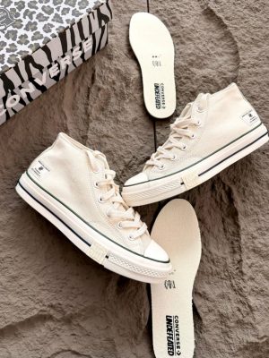 Converse x Undefeated Chuck 70 high-top sneakers in black canvas with off-white rubber sole and heel branding, held in hand with patterned shoebox and insoles visible on textured background.
