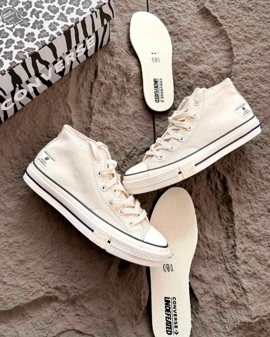 Converse x Undefeated Chuck 70 High Tops canvas Converse x Undefeated Chuck 70 high-top sneakers in black canvas with off-white rubber sole and heel branding, held in hand with patterned shoebox and insoles visible on textured background.