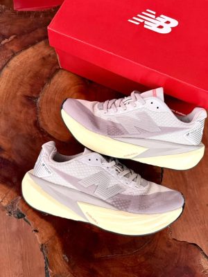 New Balance FuelCell Rebel v4 sneakers in cream and taupe mesh with gold N logo and thick white midsole, held in hand against wooden background with red NB box.
