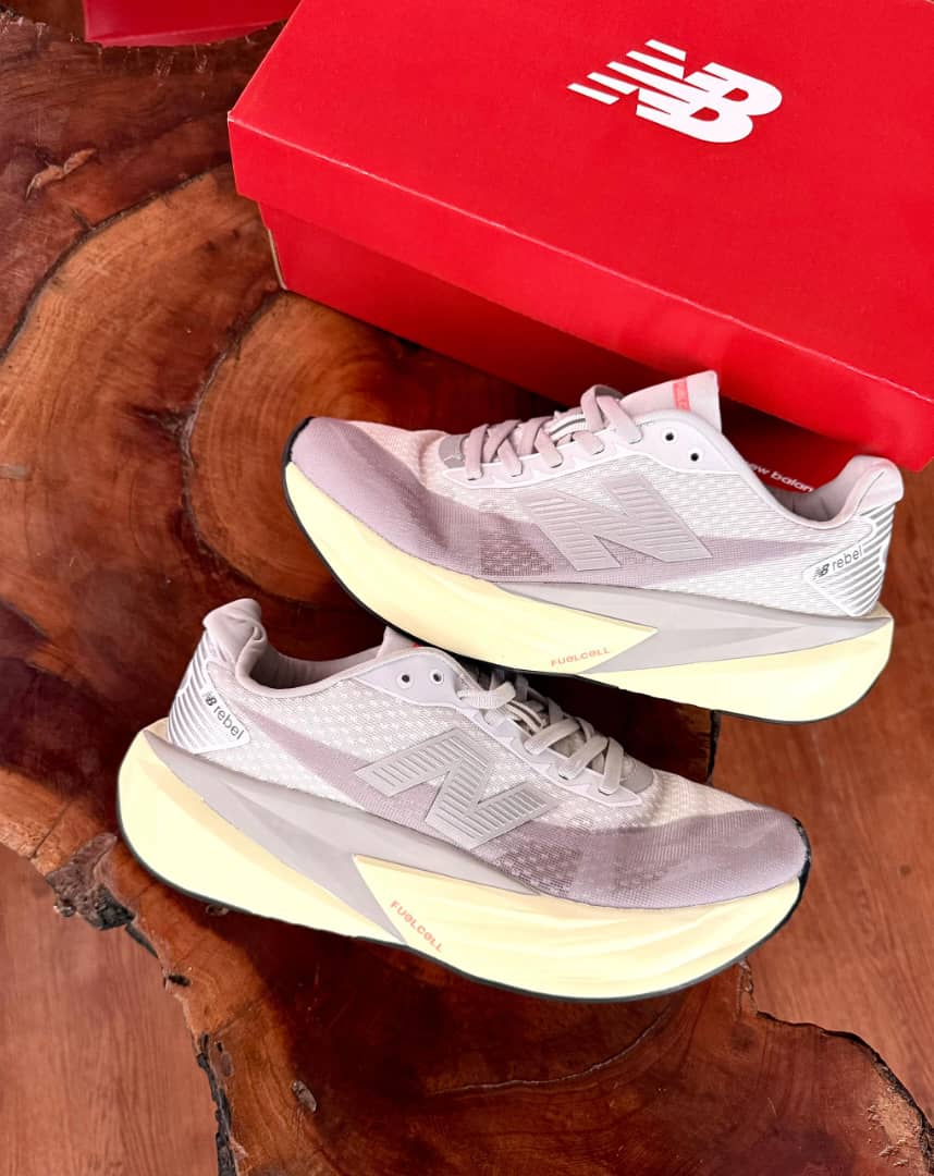 New Balance FuelCell Rebel v4 New Balance FuelCell Rebel v4 sneakers in cream and taupe mesh with gold N logo and thick white midsole, held in hand against wooden background with red NB box.