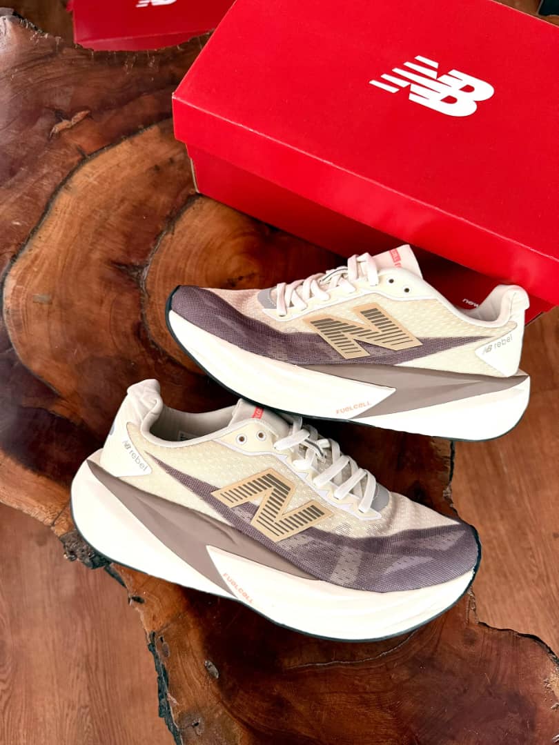New Balance FuelCell Rebel v4 sneakers in cream and taupe mesh with gold N logo and thick white midsole, held in hand against wooden background with red NB box.