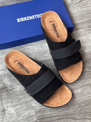 Birkenstock Arizona men's sandals in black suede with adjustable buckle straps and cork-latex footbed, displayed next to original blue Birkenstock shoebox on grey wood surface.