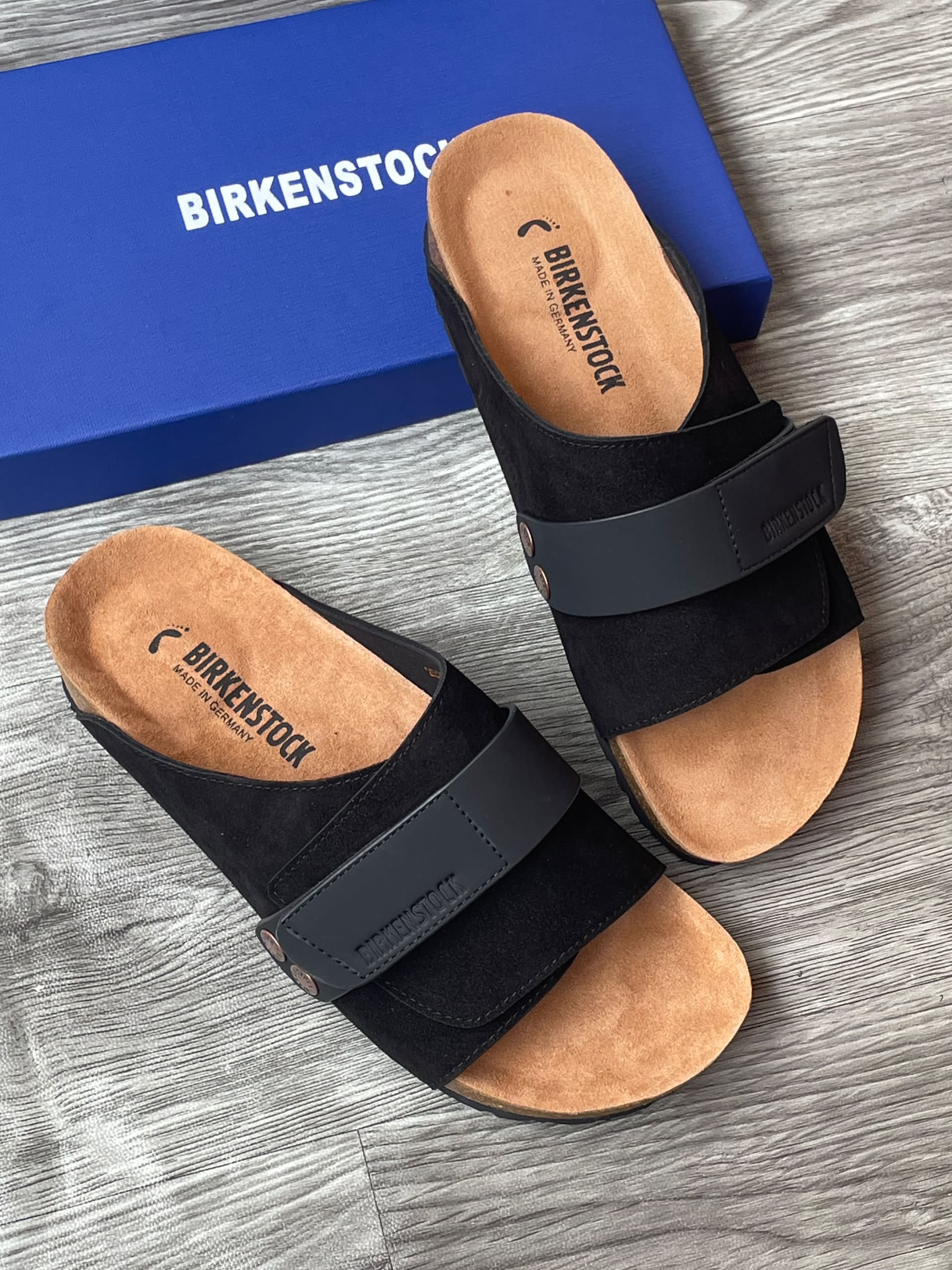 Authentic Birkenstock Arizona sandals Birkenstock Arizona men's sandals in black suede with adjustable buckle straps and cork-latex footbed, displayed next to original blue Birkenstock shoebox on grey wood surface.