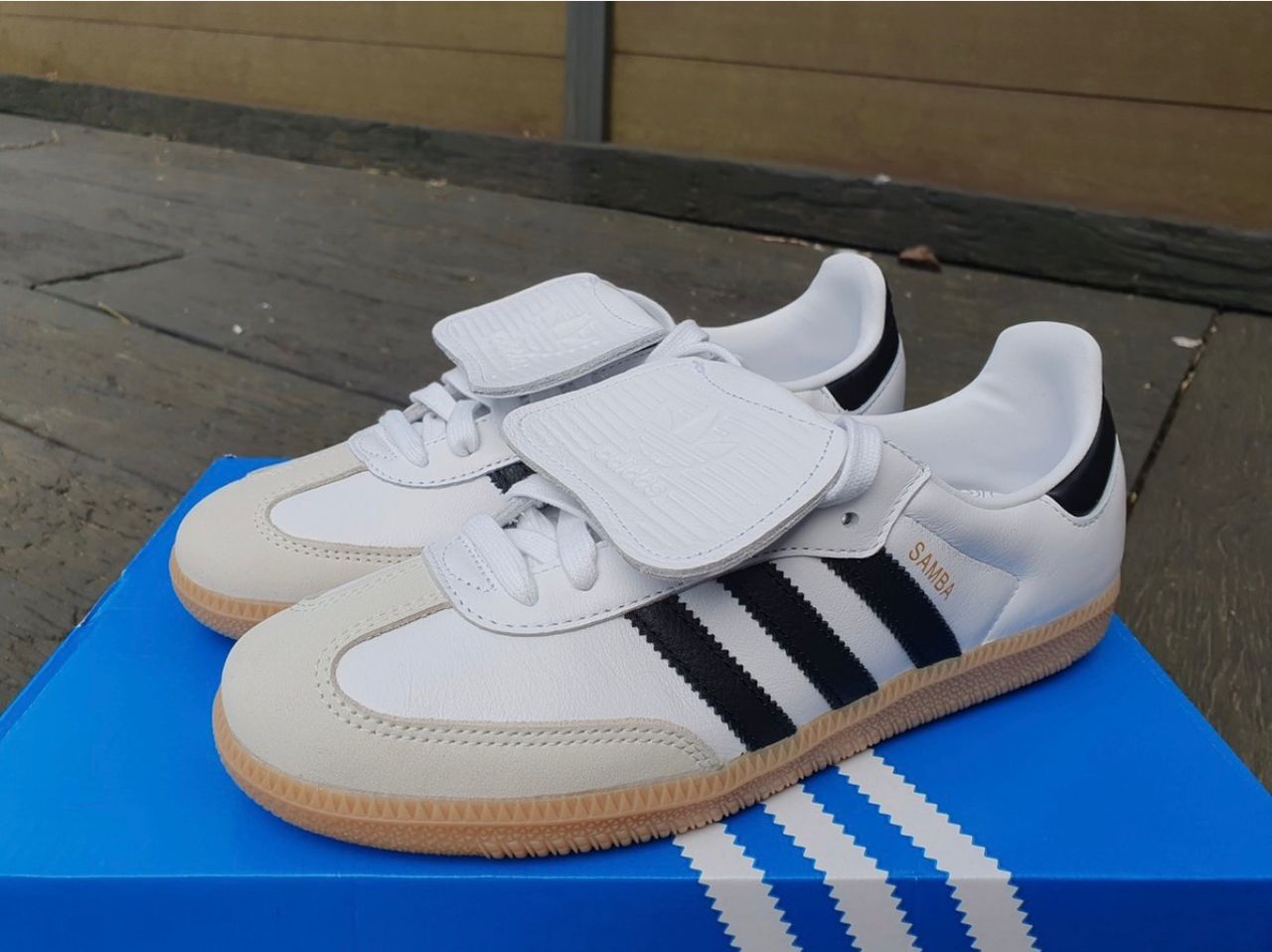 Adidas Samba OG sneakers Adidas Samba OG men's sneakers in white leather with black Three Stripes, gum rubber sole, and white velcro strap, displayed on concrete surface.