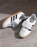 Adidas Samba OG men's sneakers in white leather with black Three Stripes, gum rubber sole, and white velcro strap, displayed on concrete surface.