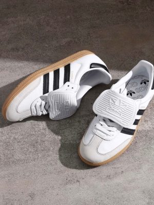 Adidas Samba OG men's sneakers in white leather with black Three Stripes, gum rubber sole, and white velcro strap, displayed on concrete surface.